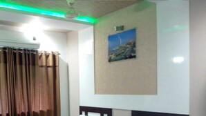 A hotel room with two beds, a ceiling fan, a painting on the wall, and a window with curtains. - Hotel Decent (Surat)