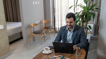 Un uomo seduto a un tavolo di legno con un portatile, una rivista e una tazza di caffè. Si vede una pianta sullo sfondo e un letto nella stanza accanto.
