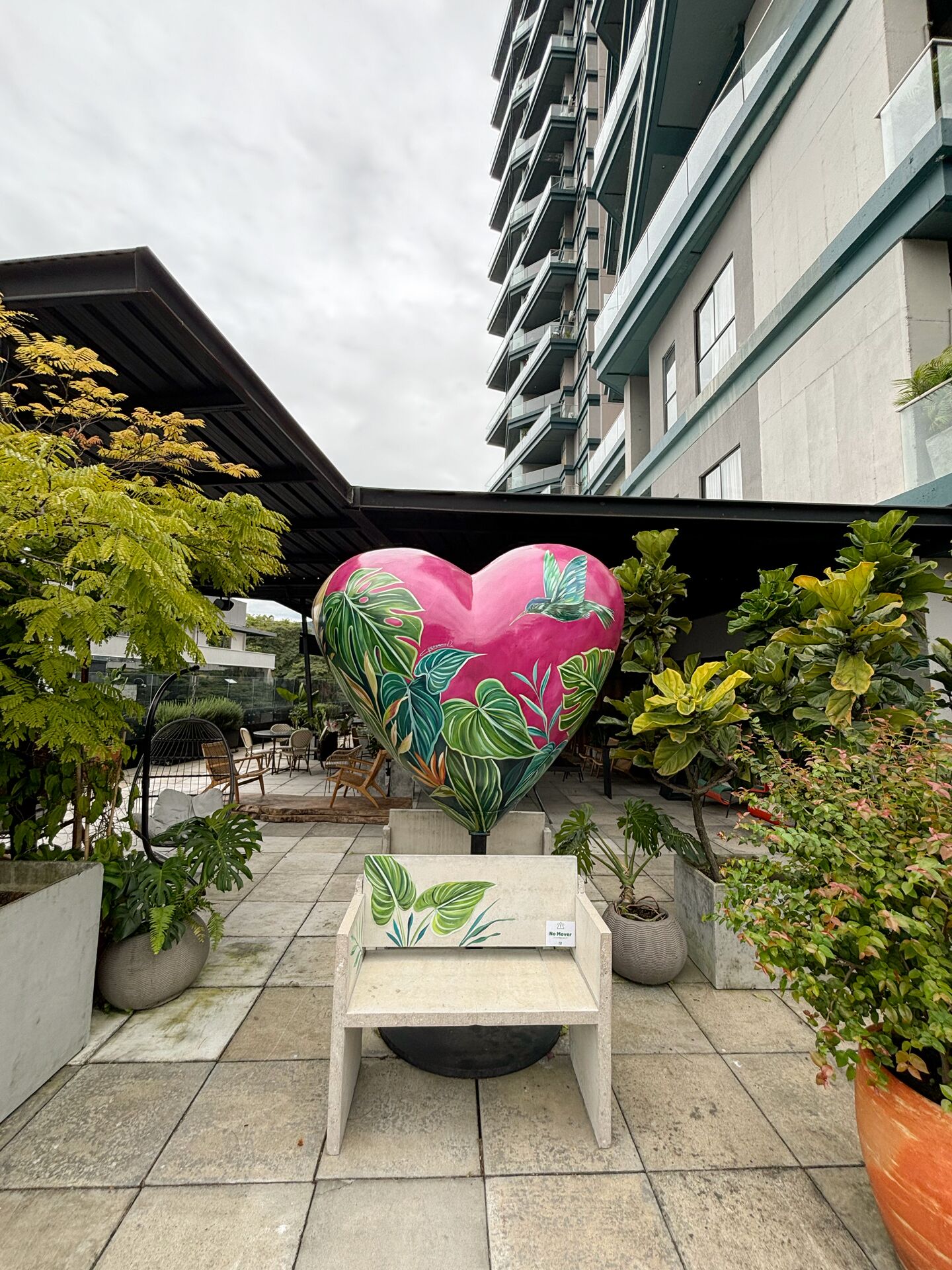 Una jardinera con forma de corazón, follaje tropical, una pequeña mesa y plantas en macetas en un patio exterior con un edificio al fondo.
