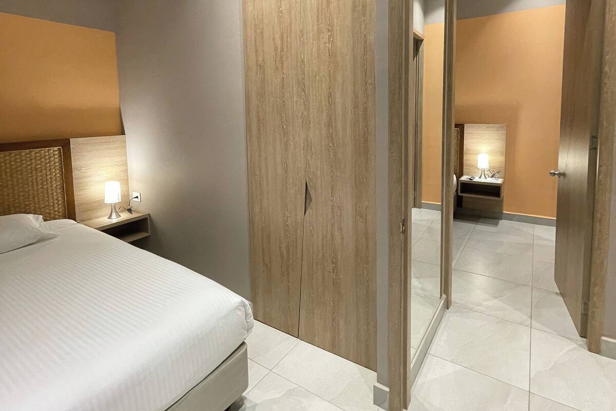 A modern hotel room with a bed, bedside table, lamp, wardrobe, and a mirror.