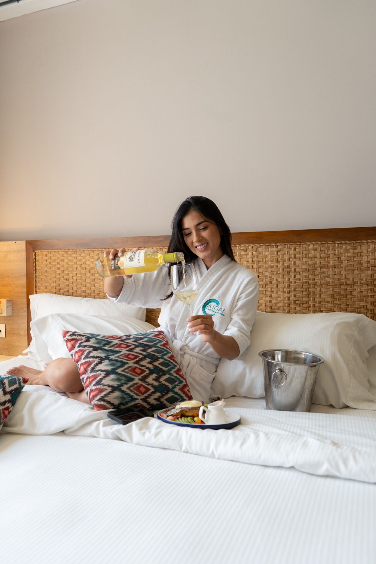 Eine Person in einem weißen Bademantel sitzt auf einem Bett mit einem Tablett Essen und einer Getränkflasche.