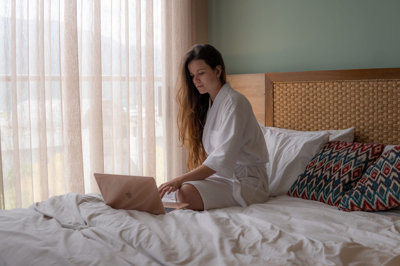 Une personne assise sur un lit, avec un ordinateur portable, un coussin à motifs et une tête de lit en osier.