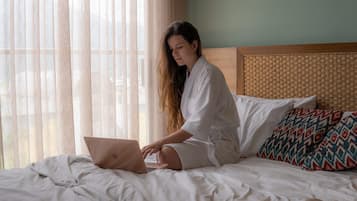 Une personne assise sur un lit, avec un ordinateur portable, un coussin à motifs et une tête de lit en osier.