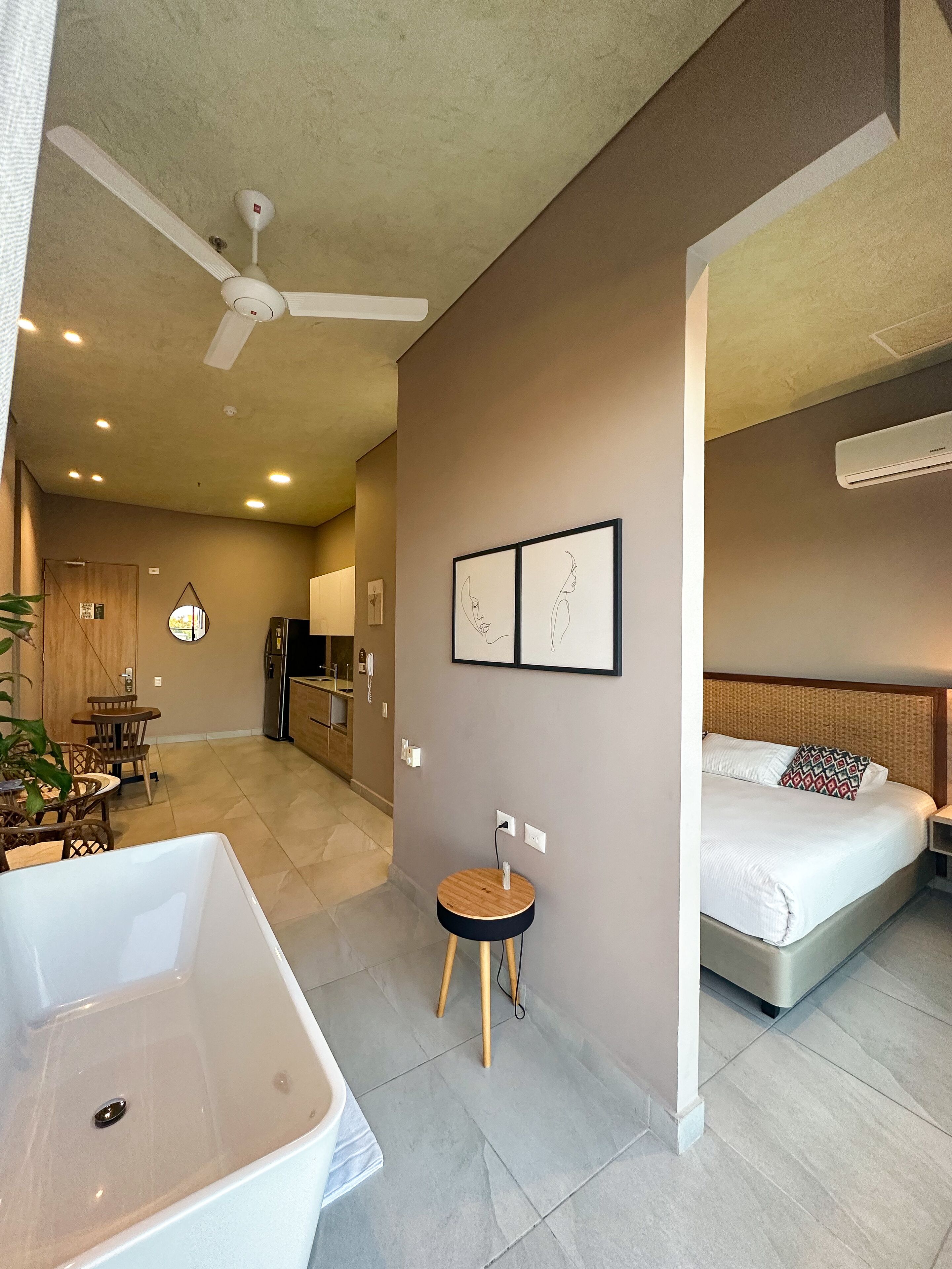 Une chambre d’hôtel moderne avec une grande baignoire, un ventilateur de plafond, un lit agrémenté d’un coussin à motifs et un petit tabouret en bois.