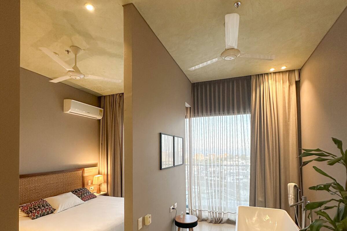 A modern hotel room with a bed, a sitting area with a table and chairs, a bathtub, and a ceiling fan.