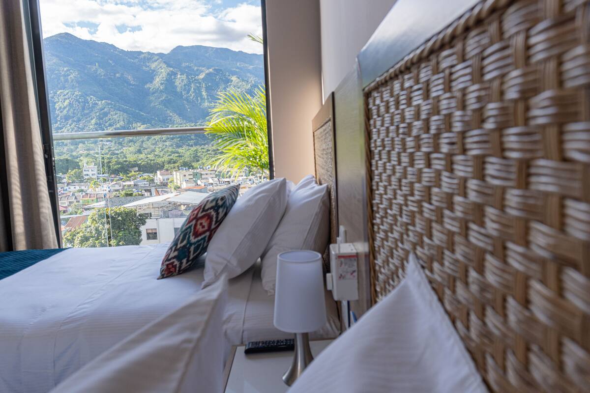 A hotel room with a bed, a nightstand, a lamp, and a view of a city and mountains.