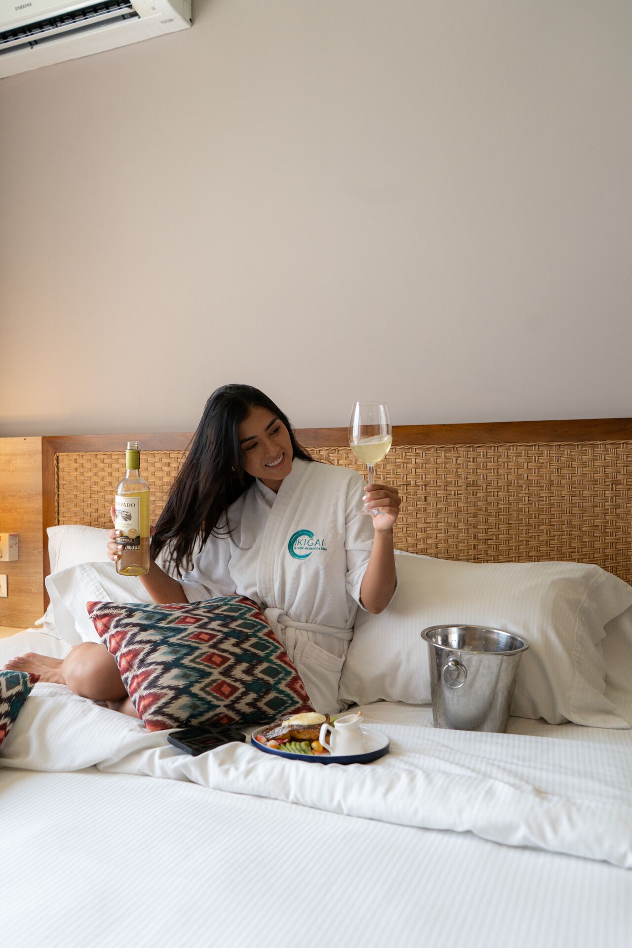 Uma pessoa sentada em uma cama com uma garrafa e um copo de vinho, um bandeja com comida e um balde de metal.