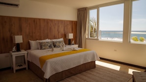 Hotelkamer met een groot bed, houten hoofdeinde, twee nachtkastjes met lampen, uitzicht op zee, en een raam met gordijnen.
