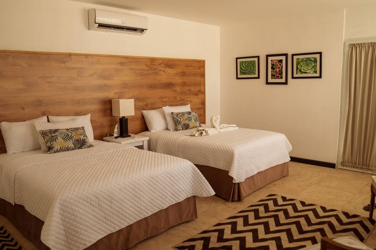 Een hotelkamer met twee bedden, een houten hoofdbord, een nachtkastje met een lamp, een raam met gordijnen en drie ingelijste foto’s aan de muur.