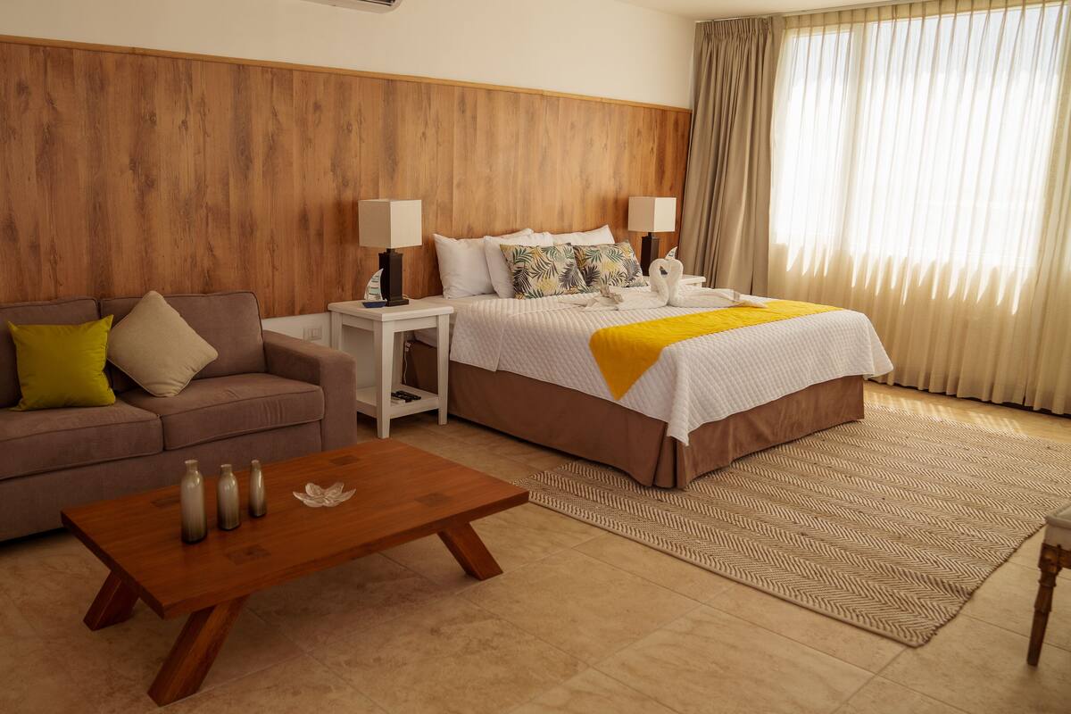 Hotelkamer met een houten hoofdbord, een bed met een wit en geel dekbed, een bruine bank met kussens, een klein wit nachtkastje met lampen en een houten salontafel.