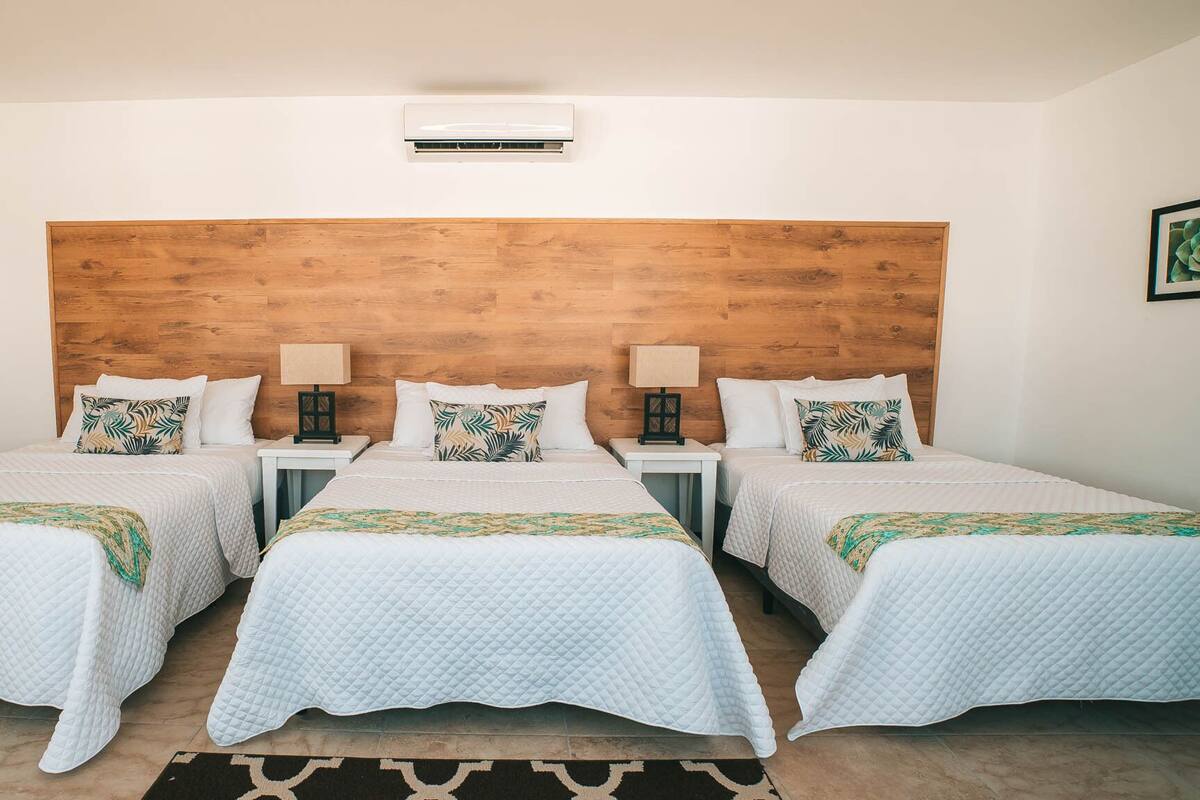 Hotelkamer met drie bedden, elk voorzien van een wit geborduurde spreideken en bijpassende kussens. Een houten hoofdbord, een klein tafeltje met een lamp en een wandgemonteerde airconditioning.