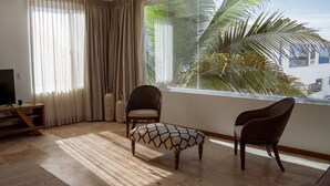 Een woonkamer met een televisie, twee stoelen, een klein poufje en een groot raam met uitzicht op palmbomen.