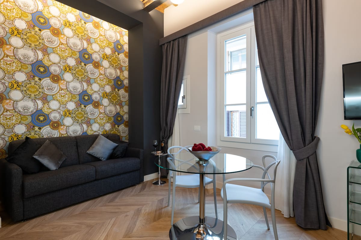 Un salón moderno con papel tapiz estampado, un sofá oscuro, una mesa de vidrio y una ventana con cortinas.