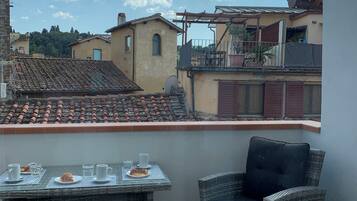 A terrace with a table set for two, a wicker chair, and a view of the surrounding buildings and sky.