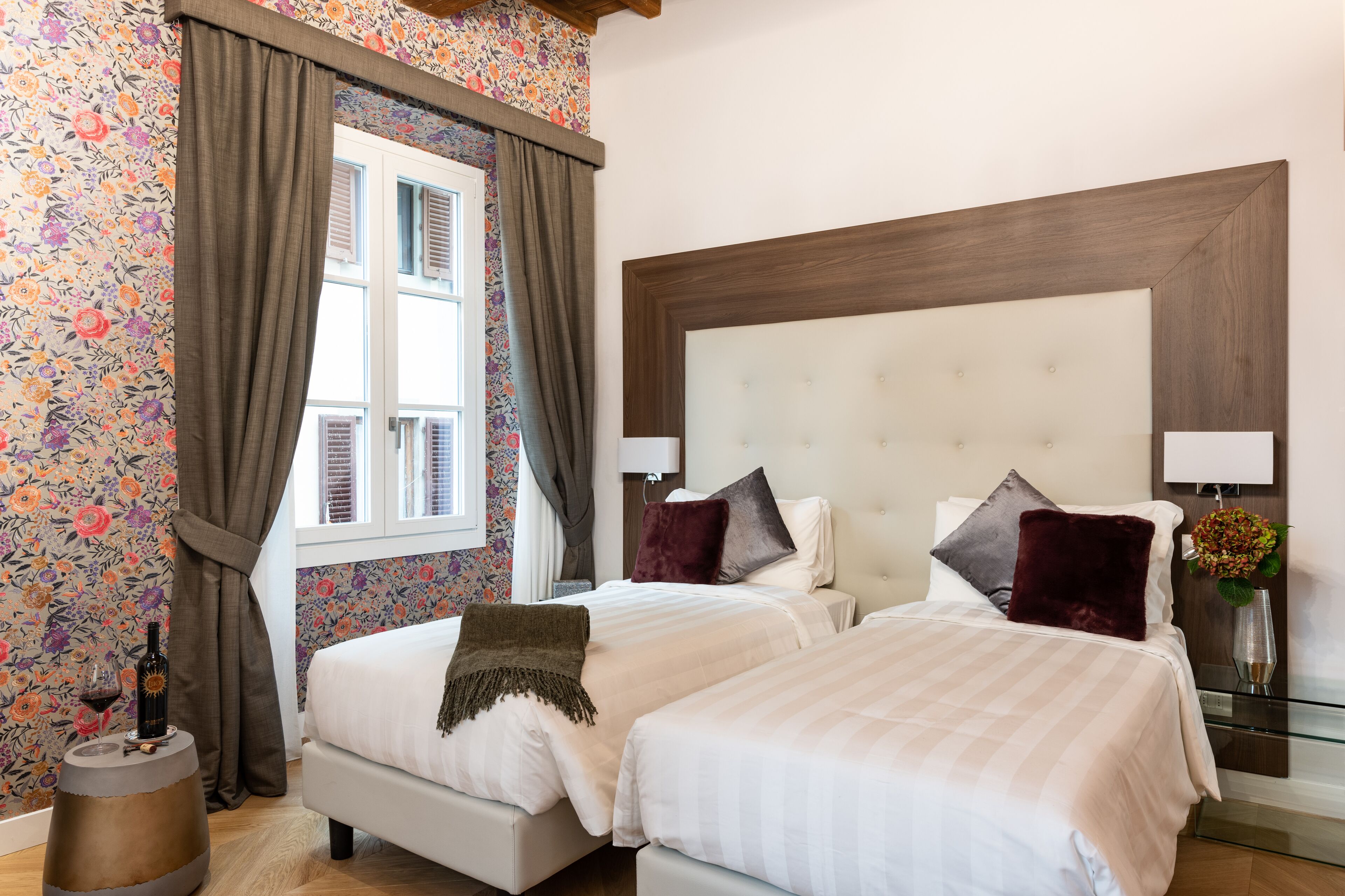 Ein Hotelzimmer mit zwei Betten, floralem Wanddesign, einem großen hölzernen Kopfteil und einem Fenster mit Vorhängen.