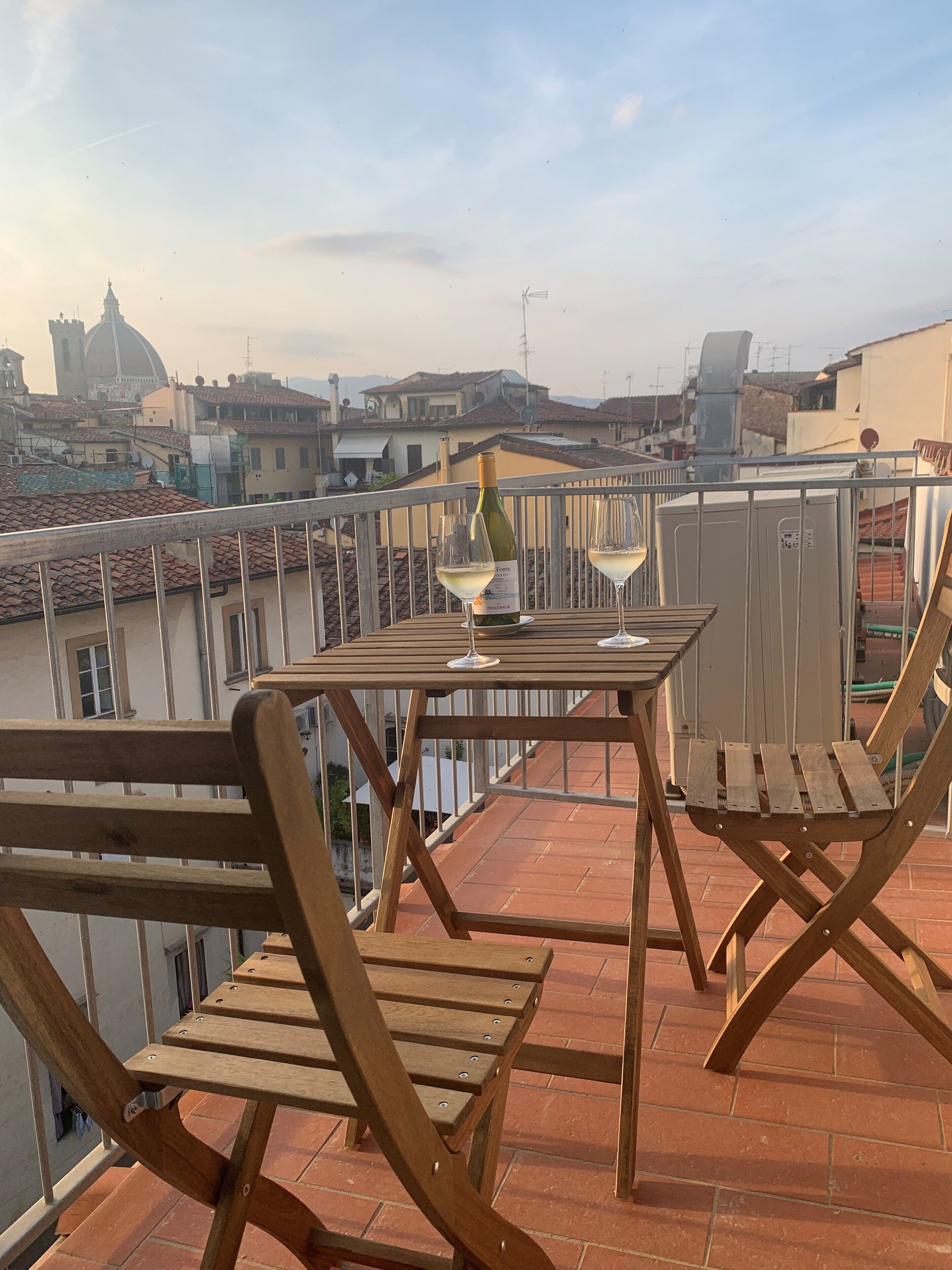Um deck de madeira com uma mesa e cadeiras, com vista para um horizonte urbano com telhados de terracota e uma estrutura em forma de cúpula em destaque ao fundo.