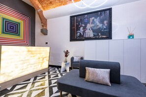 A modern lobby with a geometric patterned floor, a grey sofa with a cushion, a wall-mounted TV, and a colorful abstract painting. - Kairos by Florence Art Apartments (Florence)