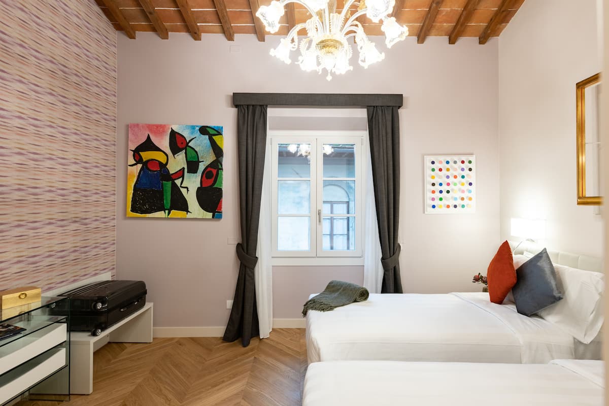 Un dormitorio con cama, escritorio, silla, ventana con cortinas, un cuadro y una lámpara de araña.