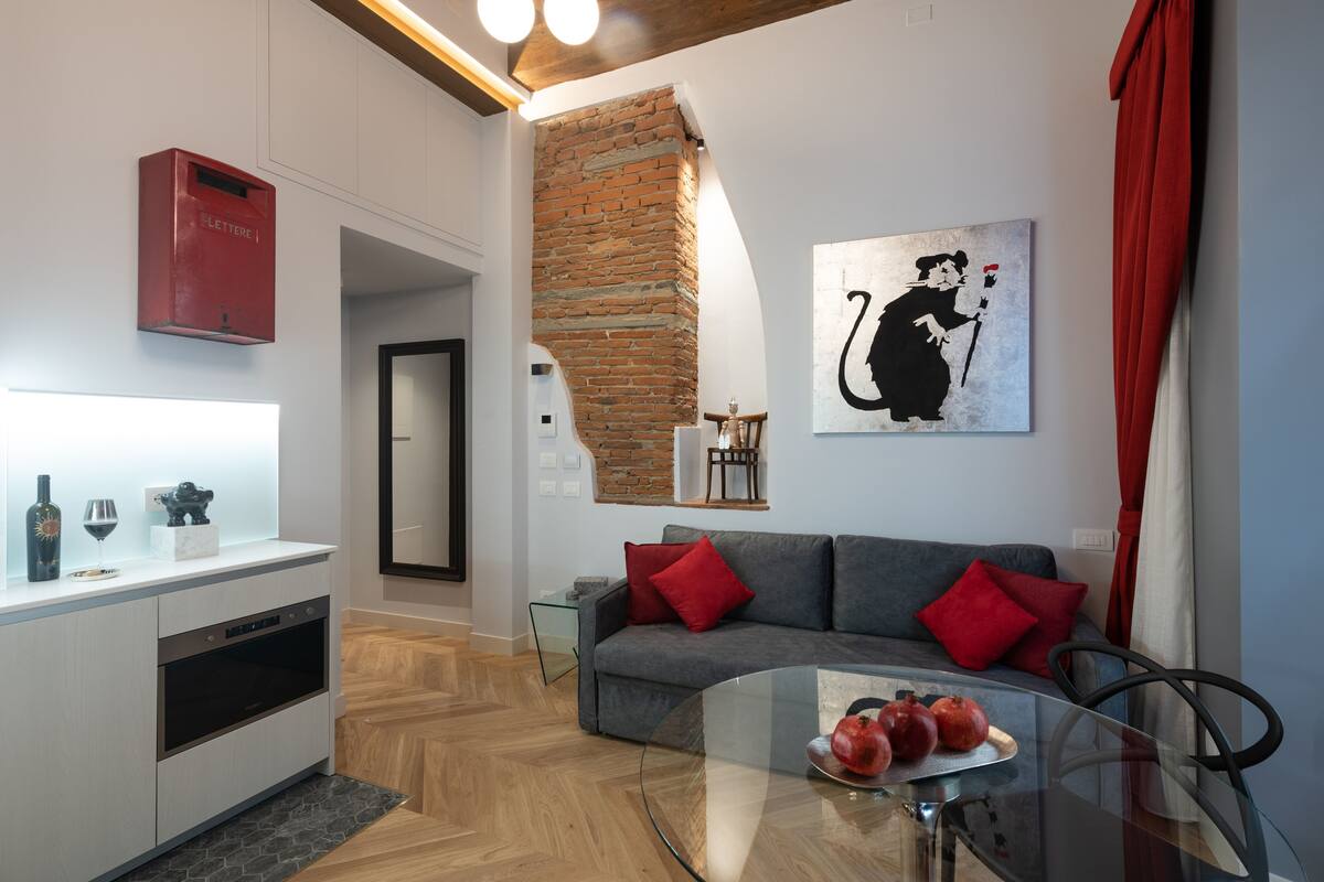 Una sala de estar moderna con un sofá gris, cojines rojos, una mesa de centro de vidrio con higos, un buzón rojo y una pared de ladrillo.