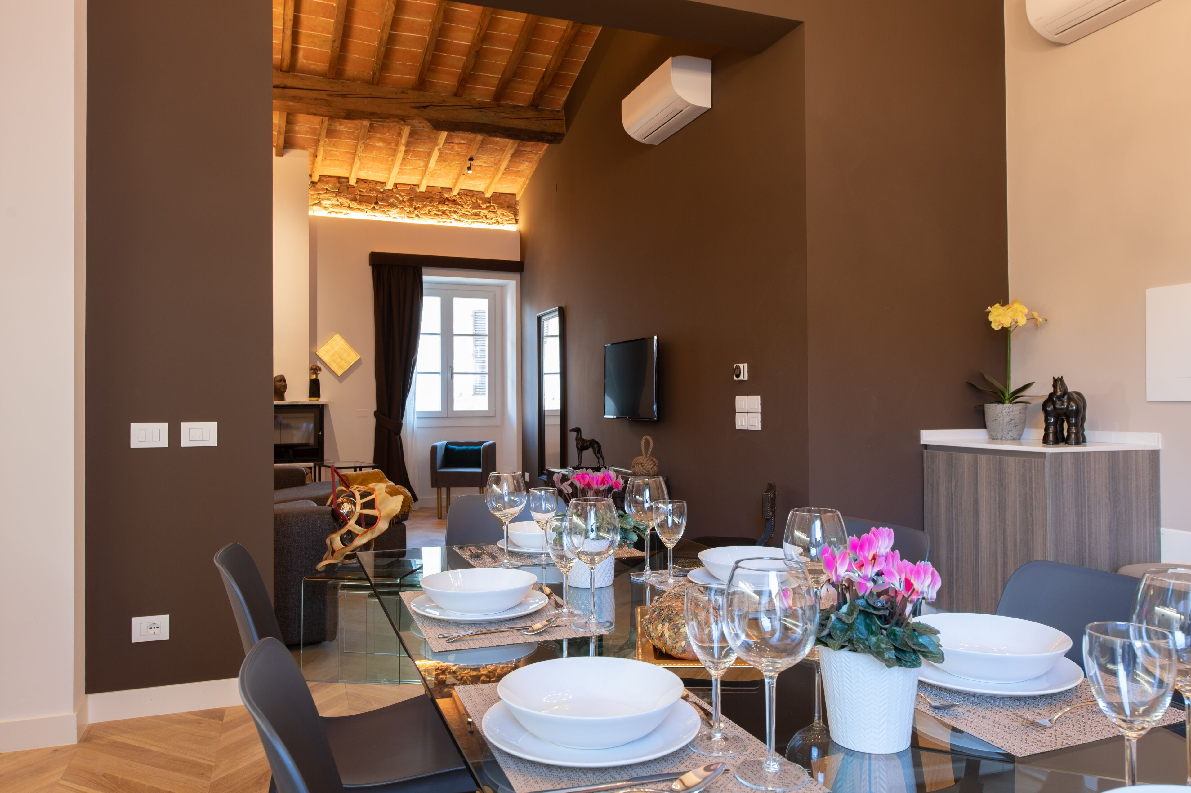 Ein Essbereich mit einem Glastisch, gedeckt für sechs Personen, umgeben von Stühlen. An der Wand ist ein Fernseher angebracht, auf einer Arbeitsplatte steht ein Blumentopf und auf dem Tisch befindet sich ein dekorativer Schädel.