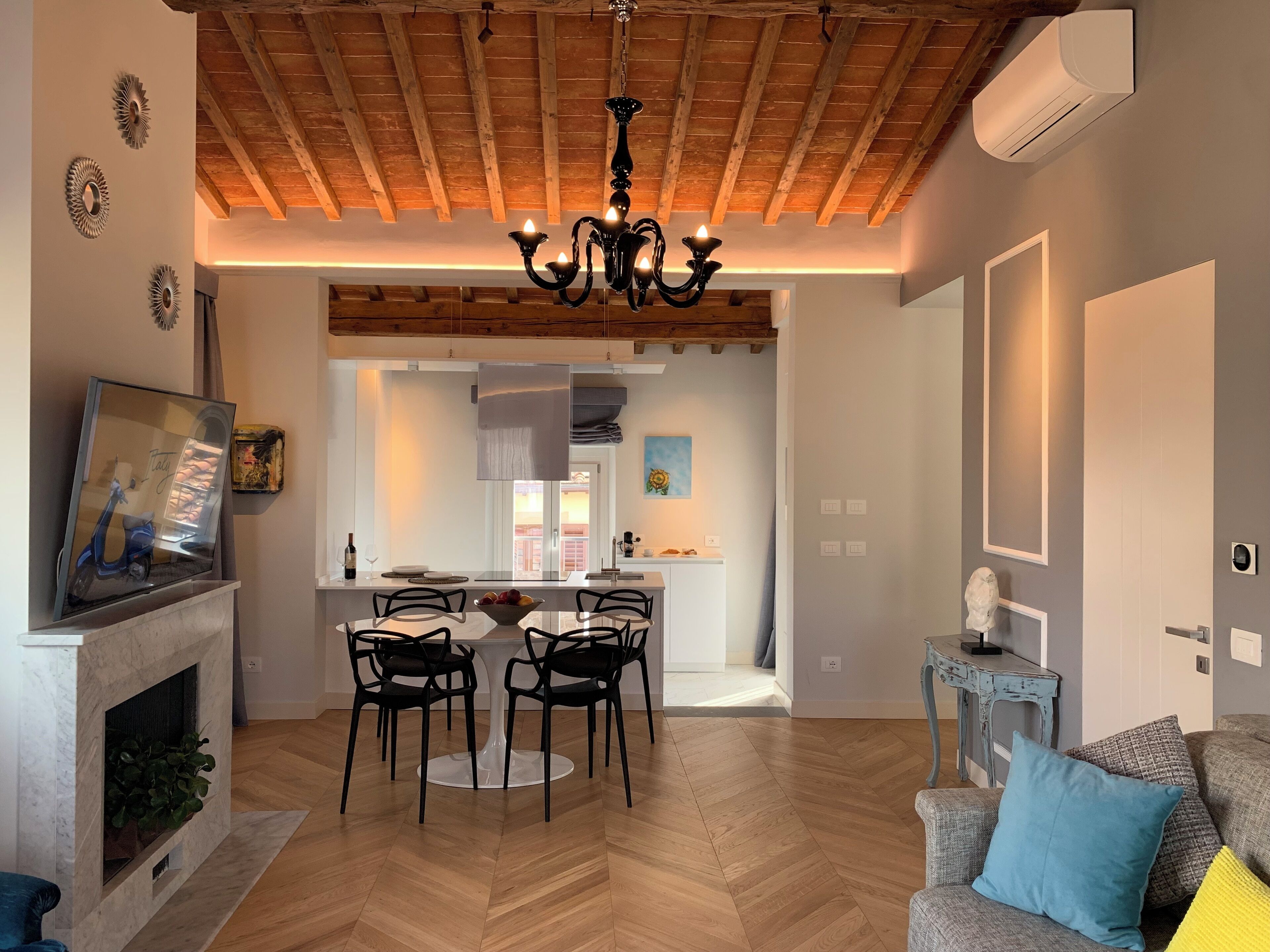A spacious living room with a herringbone wood floor, a fireplace, a dining area with a round table and chairs, and a kitchen visible through an open archway.