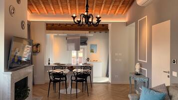 A spacious living room with a herringbone wood floor, a fireplace, a dining area with a round table and chairs, and a kitchen visible through an open archway.
