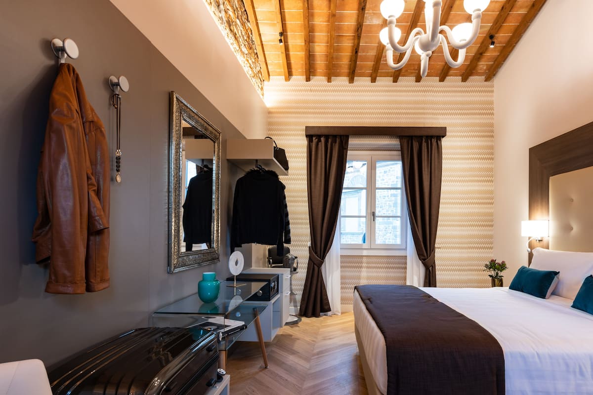 Una habitación de hotel con una cama, un escritorio, un espejo, un perchero y una ventana con cortinas.