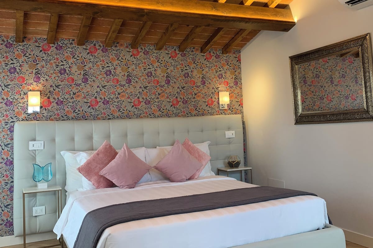 Un dormitorio con techo de madera, papel tapiz floral, cabecero capitonado, cama con ropa de cama rosa y blanca, y mesita de noche con un jarrón azul.