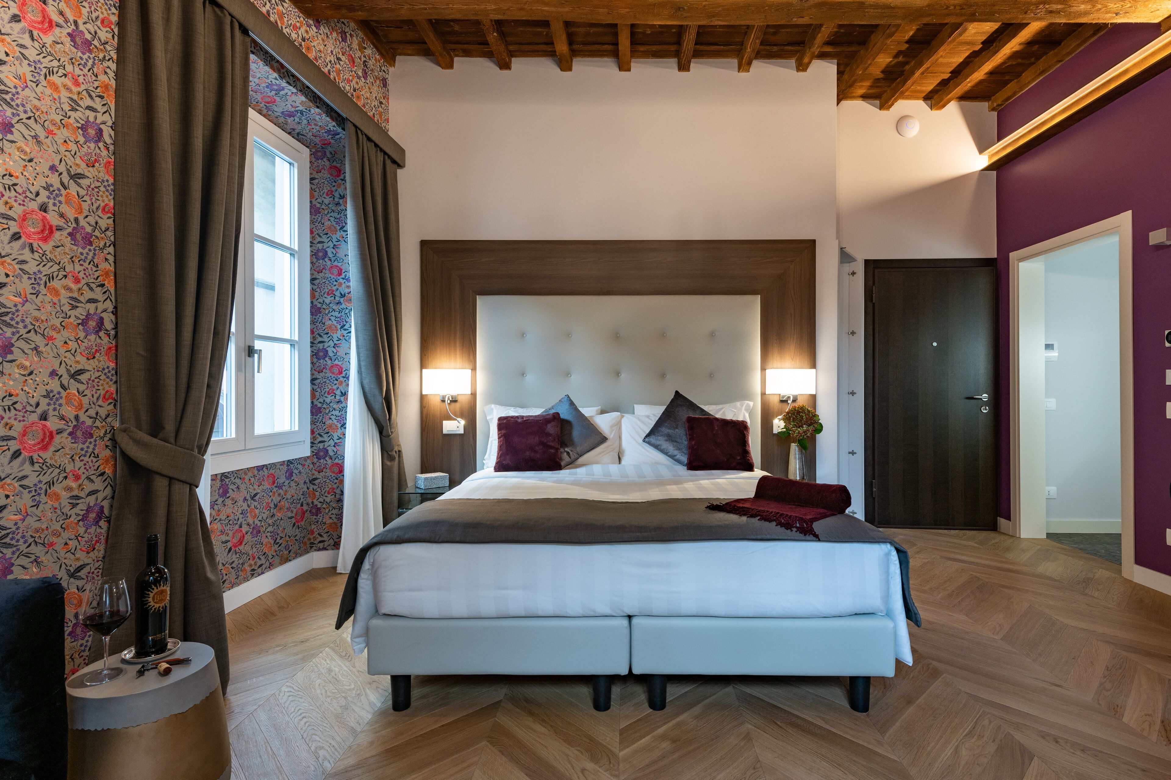A bedroom with a large bed, wooden ceiling beams, floral wallpaper, and a view of a cityscape.