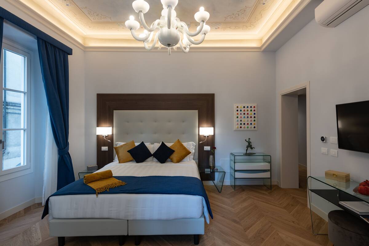 Un dormitorio moderno con una cama grande, una araña, una mesita de noche, un televisor y una obra de arte en la pared.