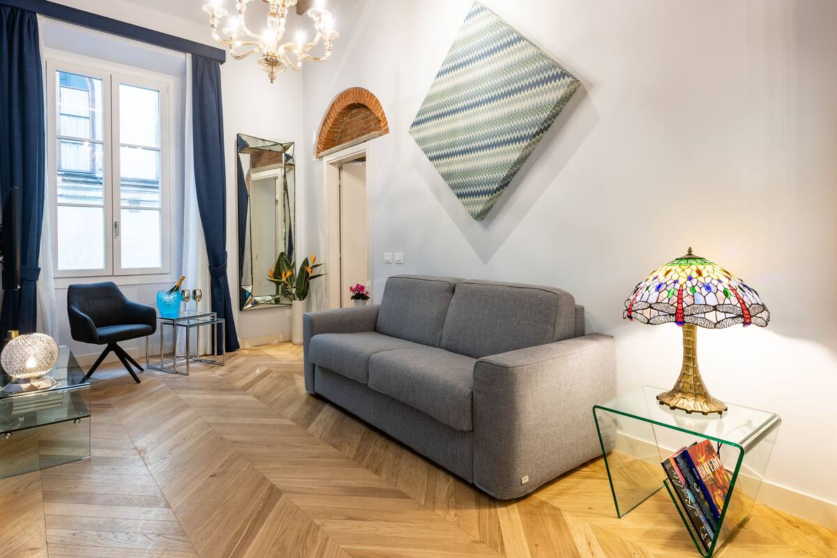 Una sala de estar moderna con un sofá gris, una mesa de centro de vidrio, una araña, un espejo montado en la pared y una lámpara decorativa.