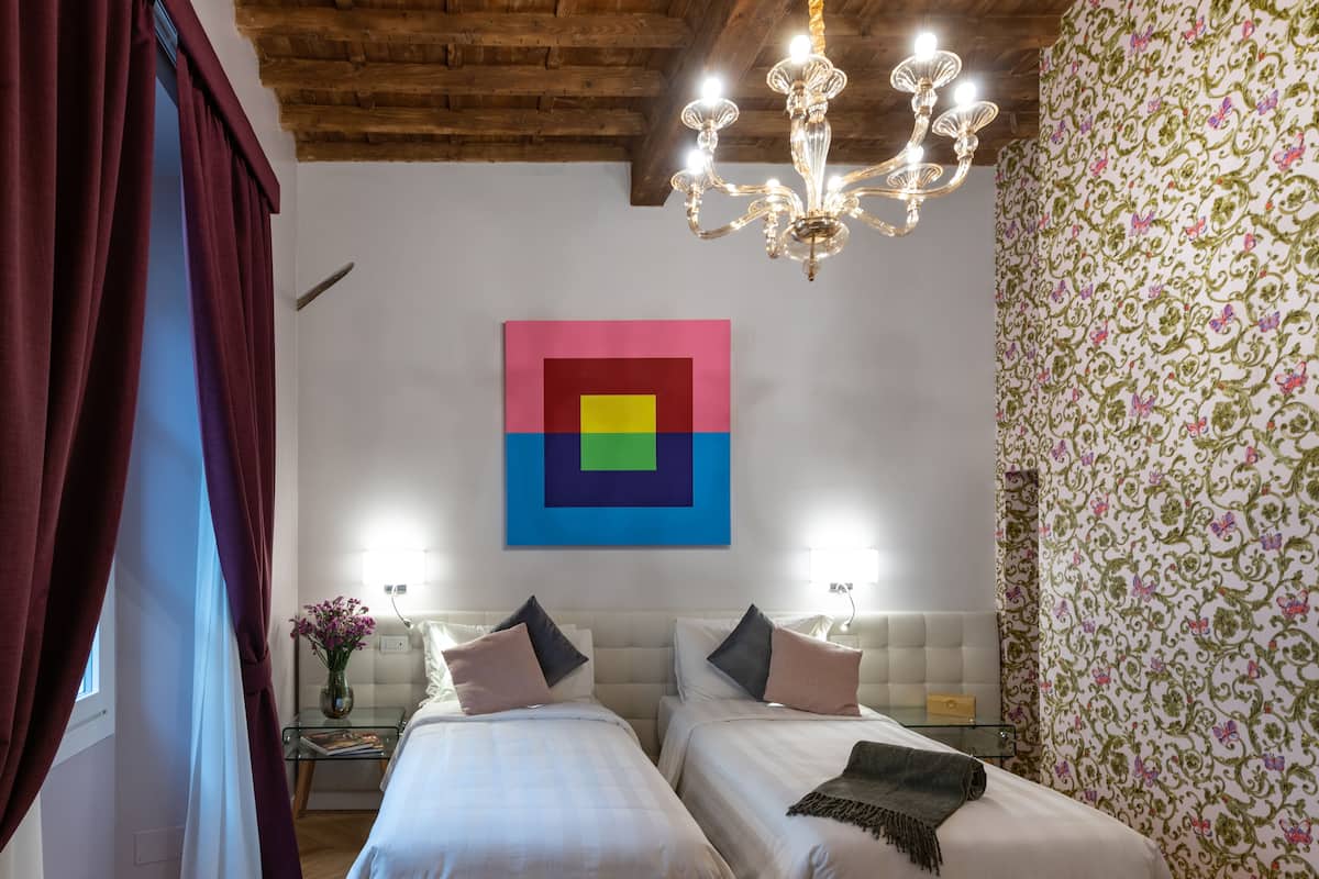 Habitación de hotel con dos camas, una araña y una obra de arte colorida, además de un papel tapiz estampado.
