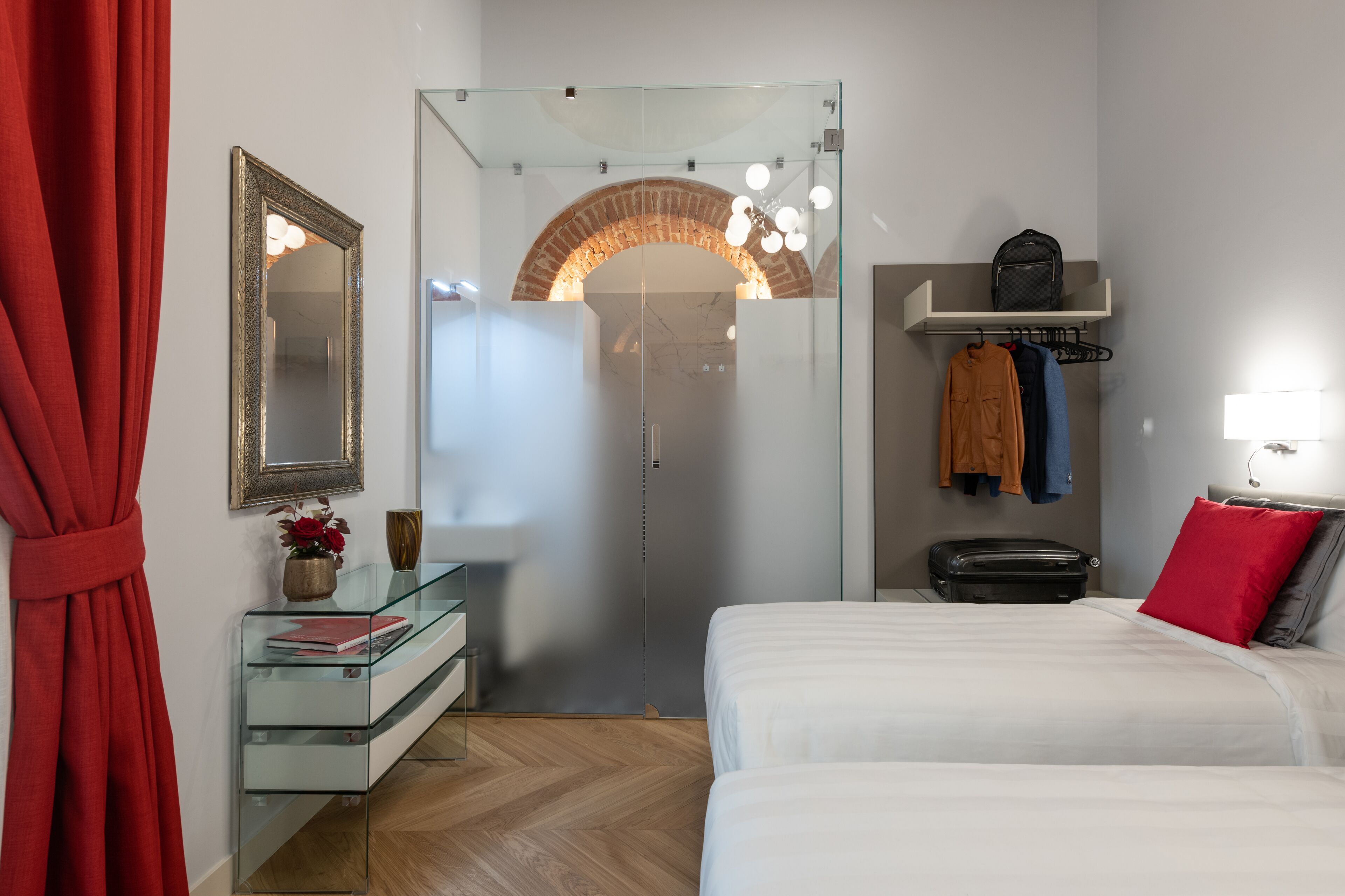 Una camera d'albergo con una scrivania in vetro, un letto con biancheria bianca, una tenda rossa, uno specchio, un arco in mattoni e un lampadario.