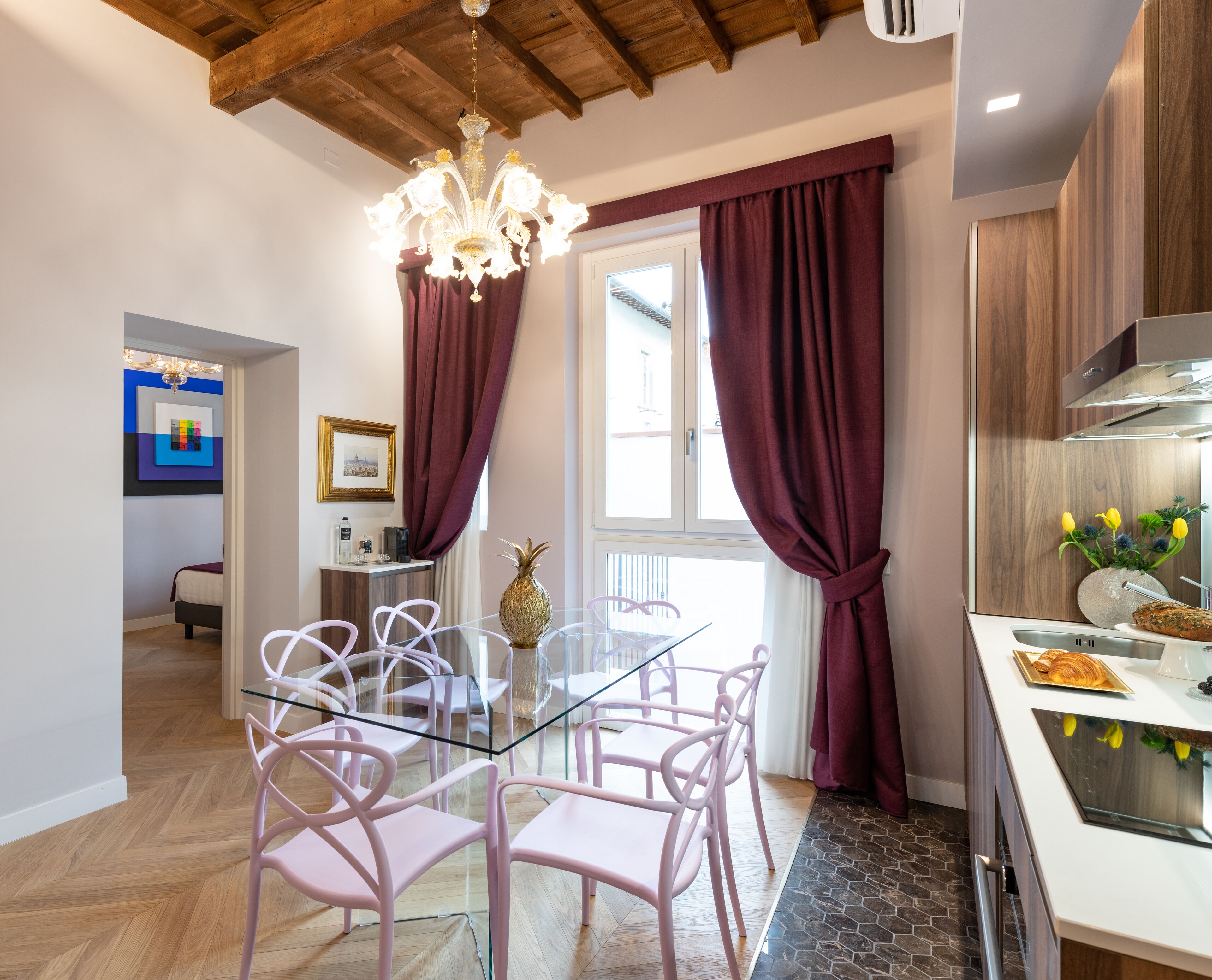 A modern kitchen with a dining area, featuring a glass table, pink chairs, and a chandelier.