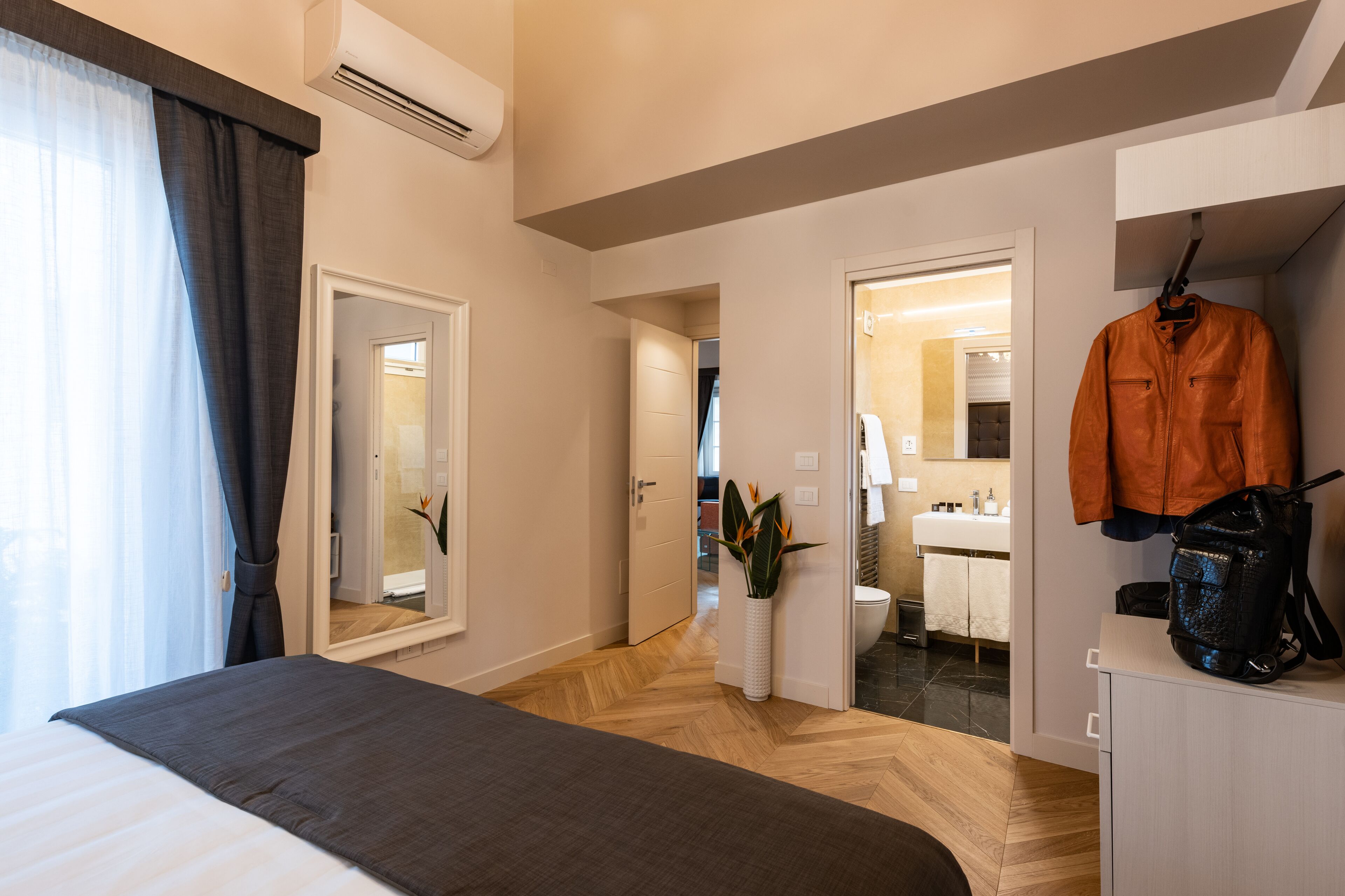 Une chambre d’hôtel avec un lit, un miroir, une plante, un porte-manteau et une salle de bain visible à travers une porte ouverte.