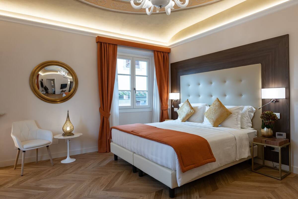 Una habitación de hotel moderna con una cama grande, un espejo redondo, una mesita de noche con una lámpara y una silla.