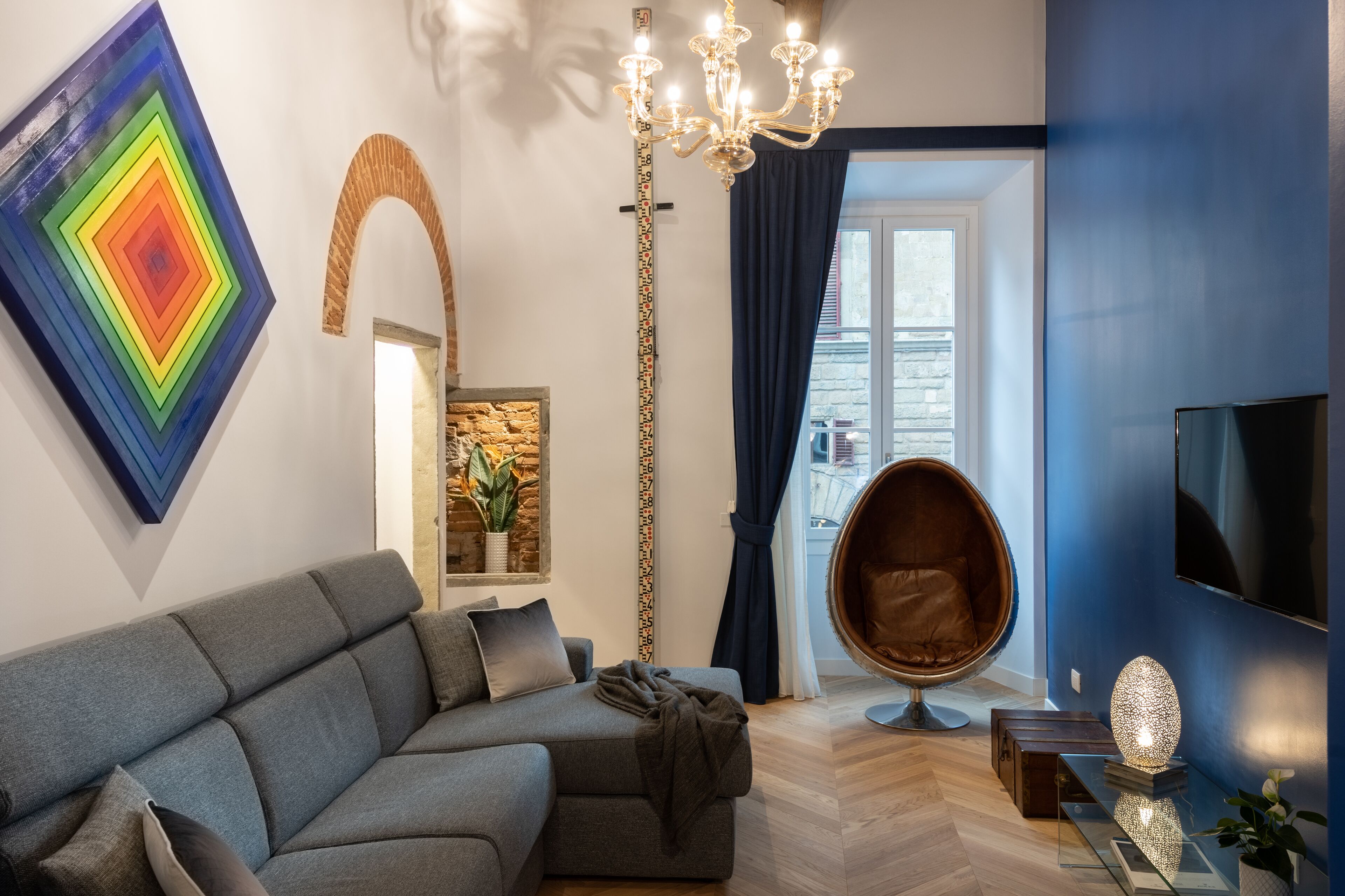 A modern living room with a grey sofa, a tall decorative mirror, a chandelier, and a large TV on the wall.