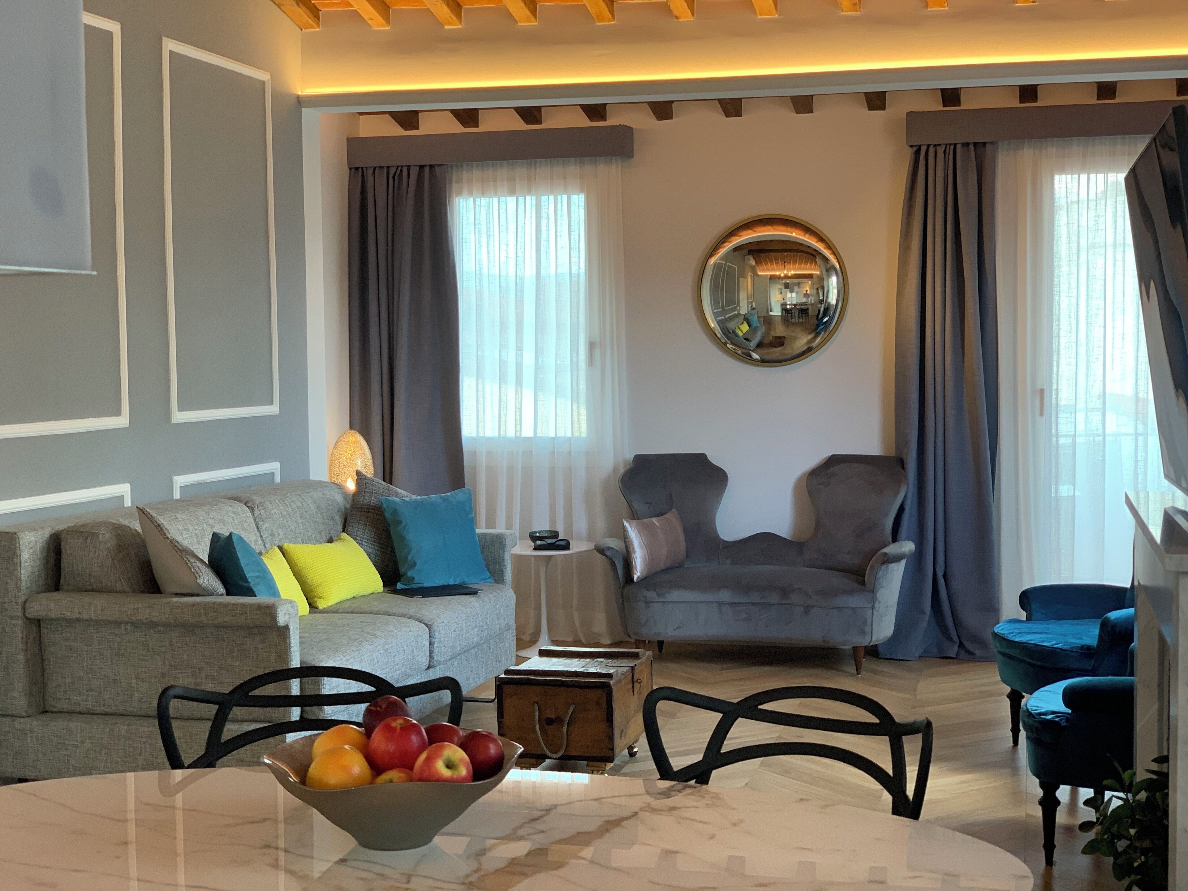 Een moderne woonkamer met een grijze bank, een blauwe fauteuil, een ronde spiegel en een tafel met een fruitschaal.