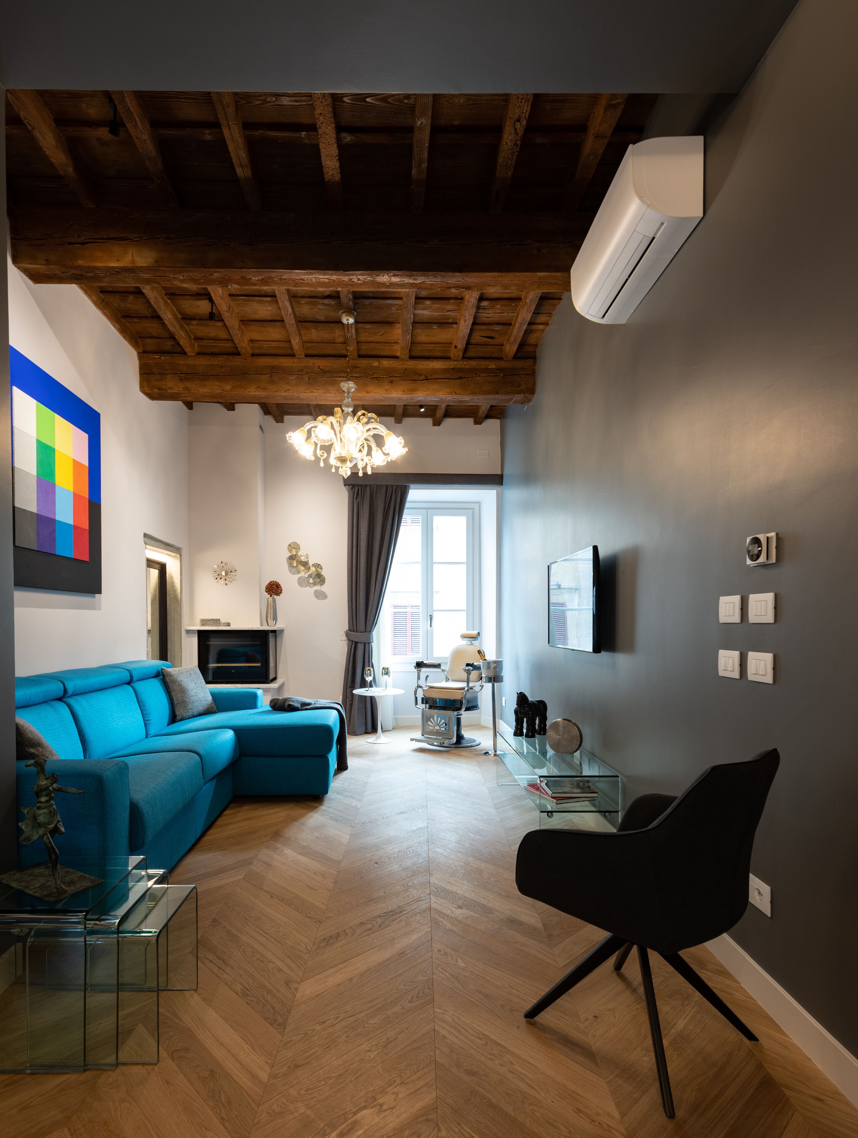 Una sala de estar moderna con un sofá seccional azul, una mesa de centro de vidrio, una silla negra y un piso de madera con patrón de espiga.