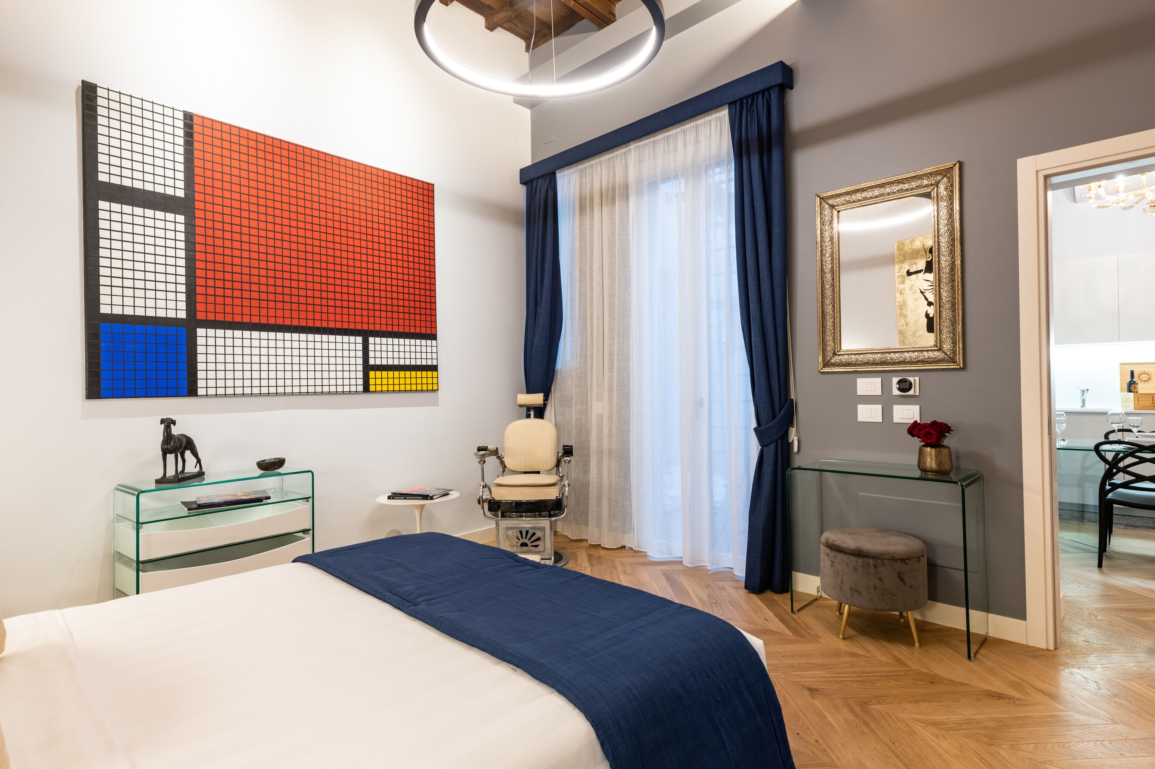 A modern bedroom with a bed, a glass desk, a chair, a mirror, and a large abstract art piece on the wall.