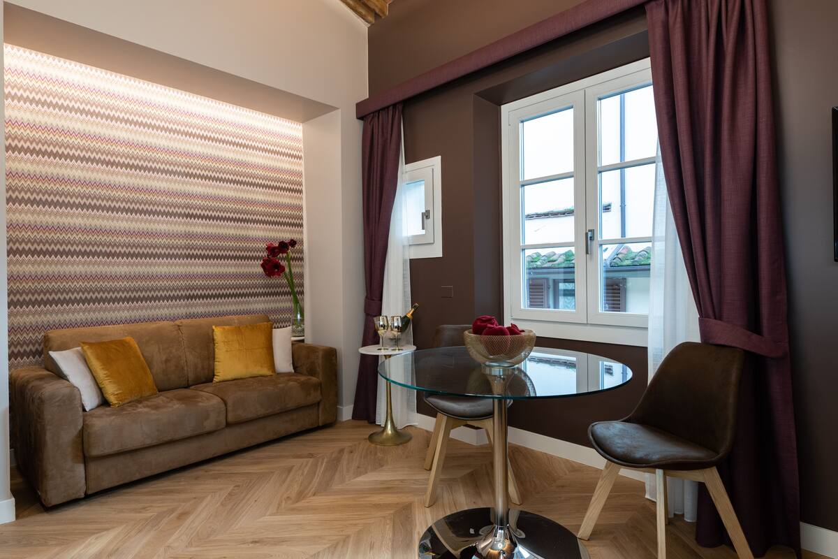 Una sala de estar moderna con una mesa de centro de vidrio, un sofá marrón con cojines, un sillón oscuro y una ventana con cortinas.