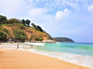 A beach with clear turquoise water, a sandy shore, and a hilly backdrop with trees.