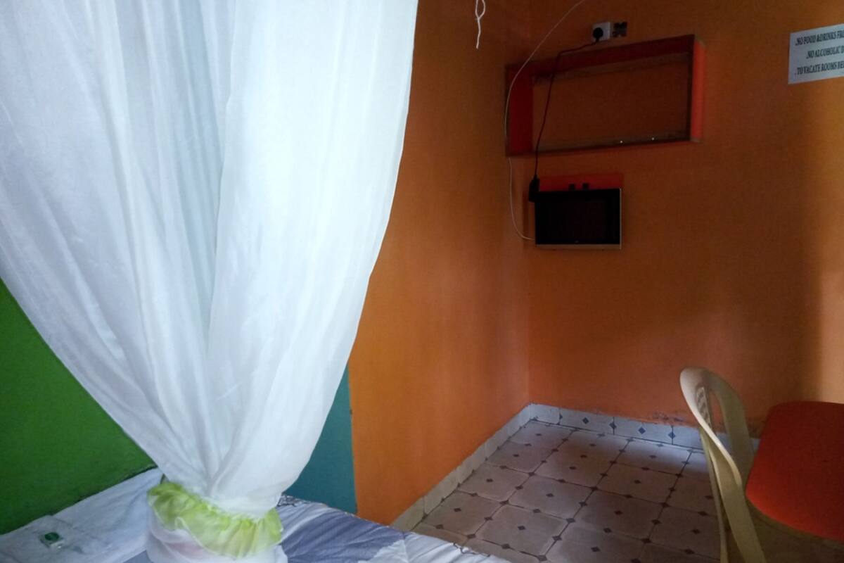A small, simple room with a bed, a chair, and a table. There is a sign on the wall and a power outlet above the bed.