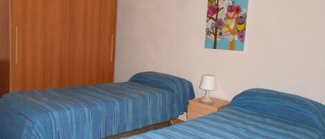 A room with two beds, a wooden wardrobe, a nightstand with a lamp, and a painting on the wall.