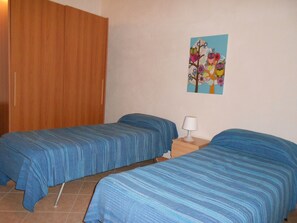 Een kamer met twee bedden, een houten garderobe, een nachtkastje met een lamp en een schilderij aan de muur.