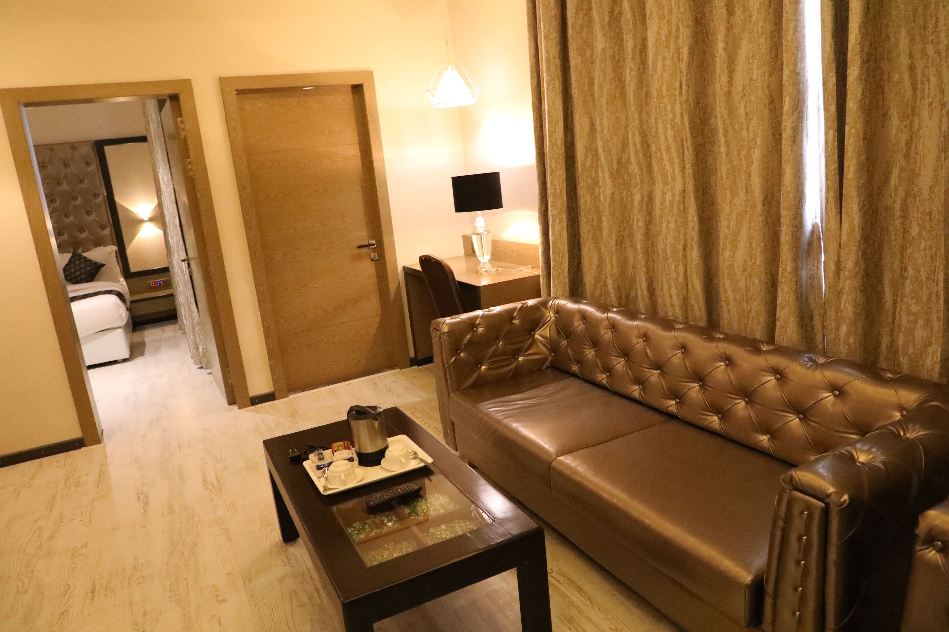 A hotel room with a brown leather sofa, a wooden coffee table with a tray of items, a lamp on a side table, and a door leading to a bathroom.
