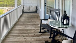 Une véranda couverte avec un plancher en bois, des balustrades blanches, une balançoire, une table avec des chaises et une vue sur la verdure.