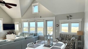 Amplia sala de estar con un gran ventanal con vista a la playa, una mesa de comedor con individuales y un ventilador de techo.