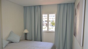 Quarto com cama, mesa de cabeceira, abajur e janela com cortinas.