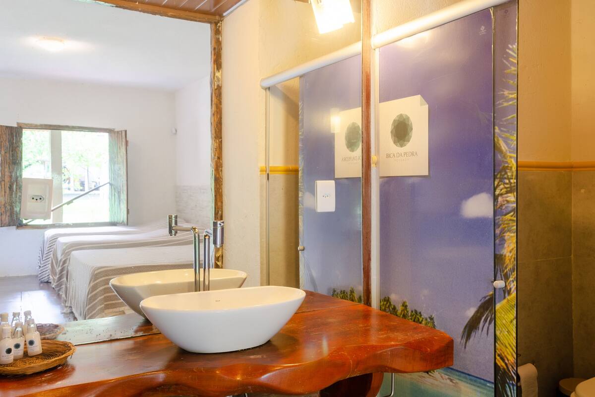 Une salle de bain avec un grand miroir, un lavabo blanc et un meuble-lavabo en bois. On aperçoit deux lits au fond de la pièce.